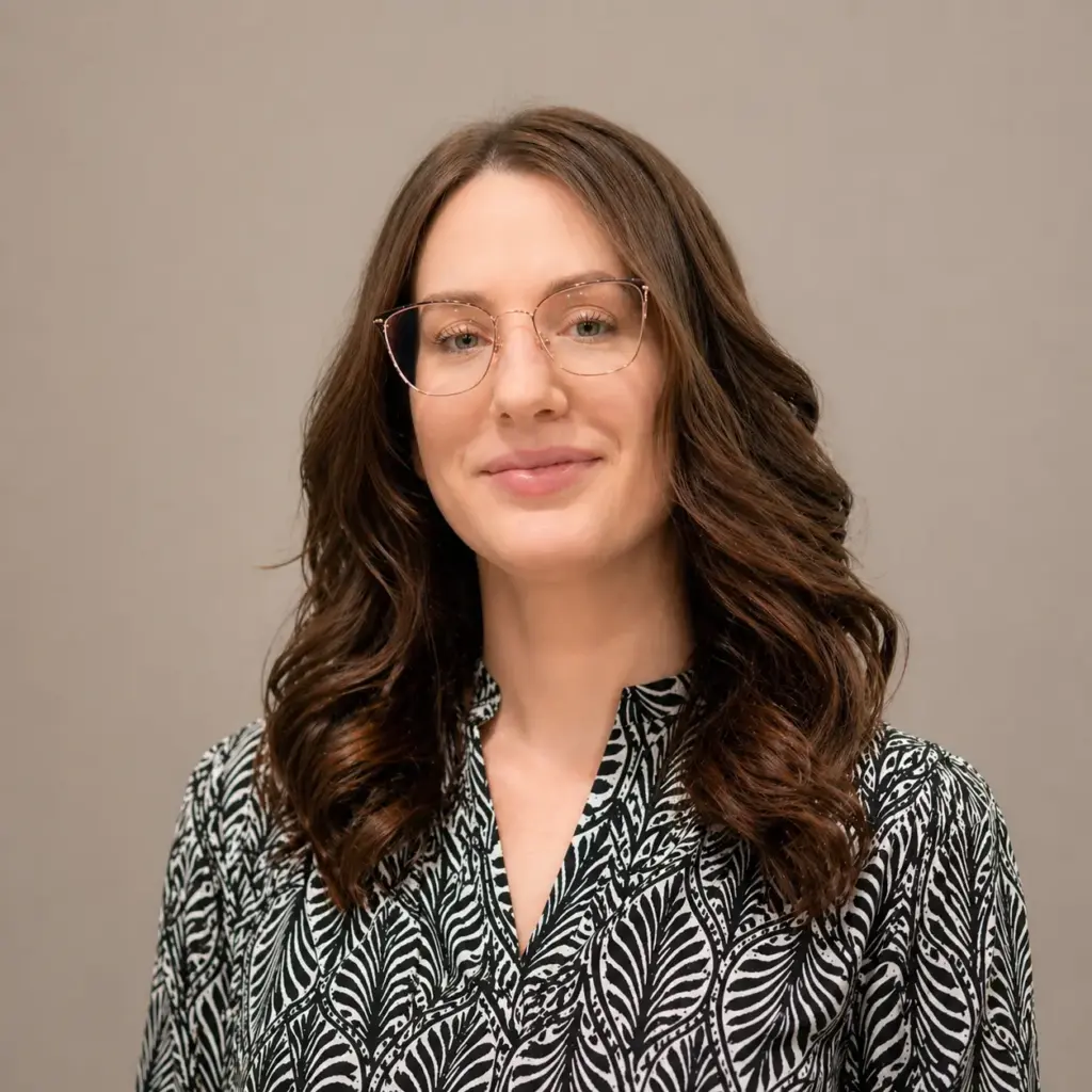 Portrait of a smiling woman with wavy brown hair and round glasses, wearing a black-and-white patterned blouse against a neutral background.