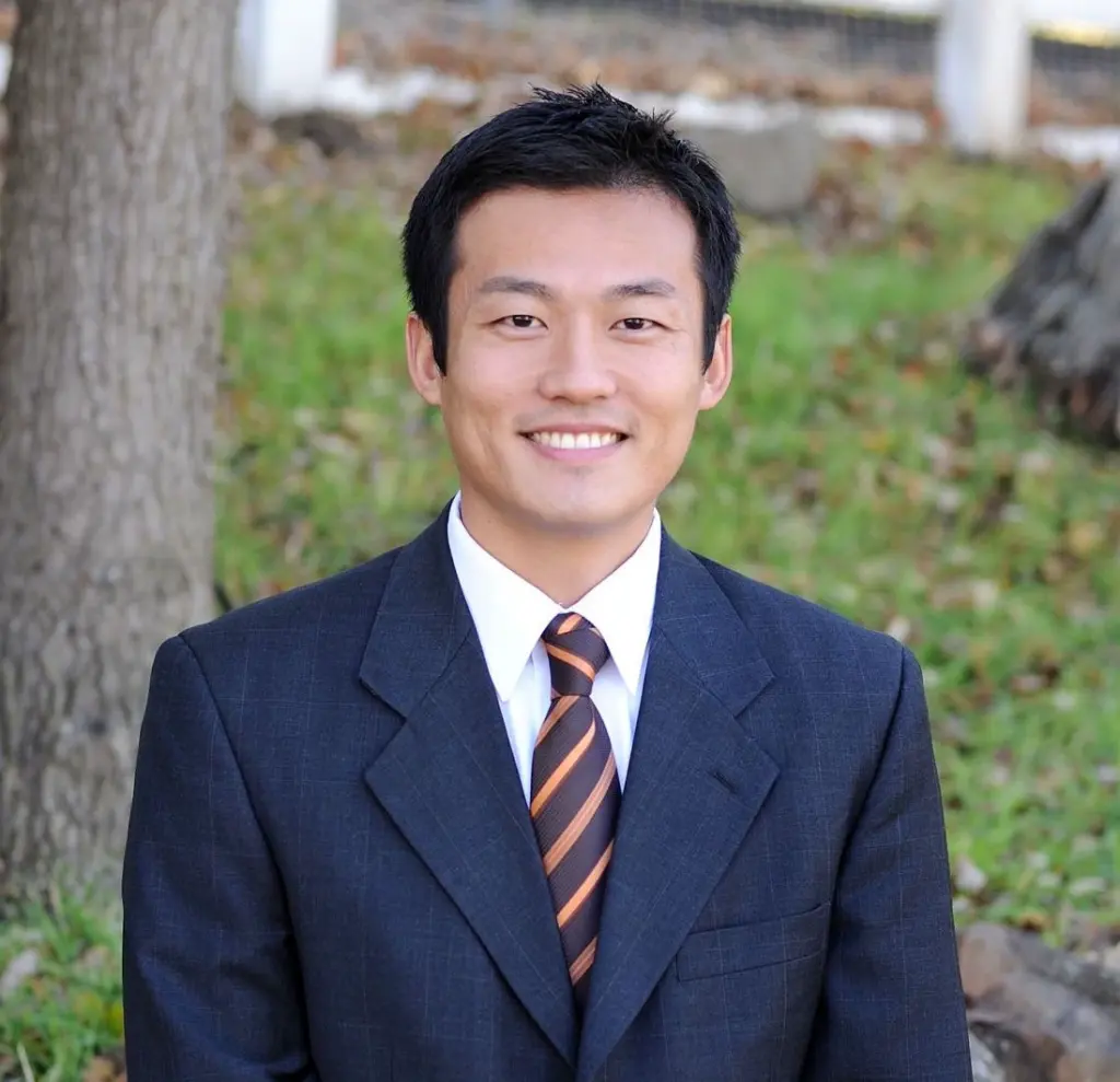 Smiling man in a dark suit and striped tie outdoors, with trees and grass in the background.